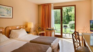 Habitación de hotel con dos camas, un escritorio con silla, una mesita con una silla y un gran ventanal corredero que da a un espacio exterior con césped y una mesa.