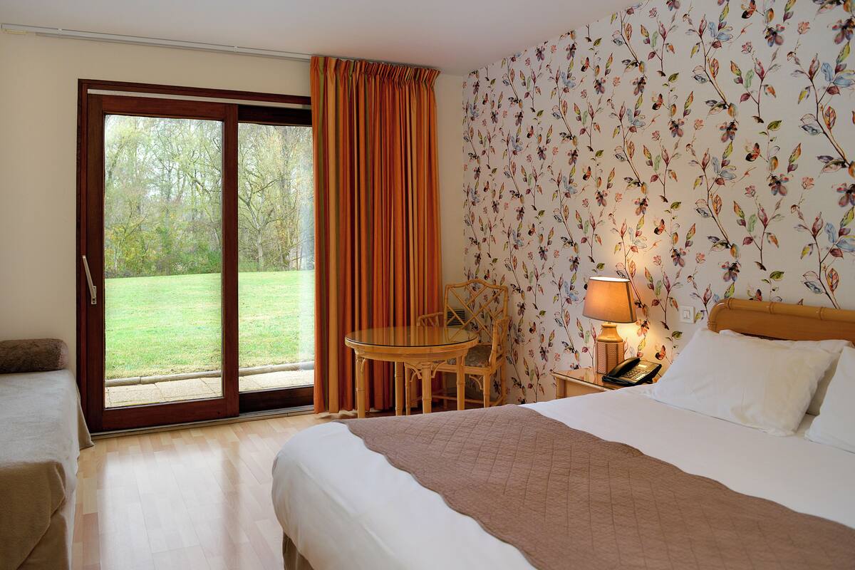 A hotel room with a bed, a table, a chair, a lamp, and a view of a garden through a sliding door.