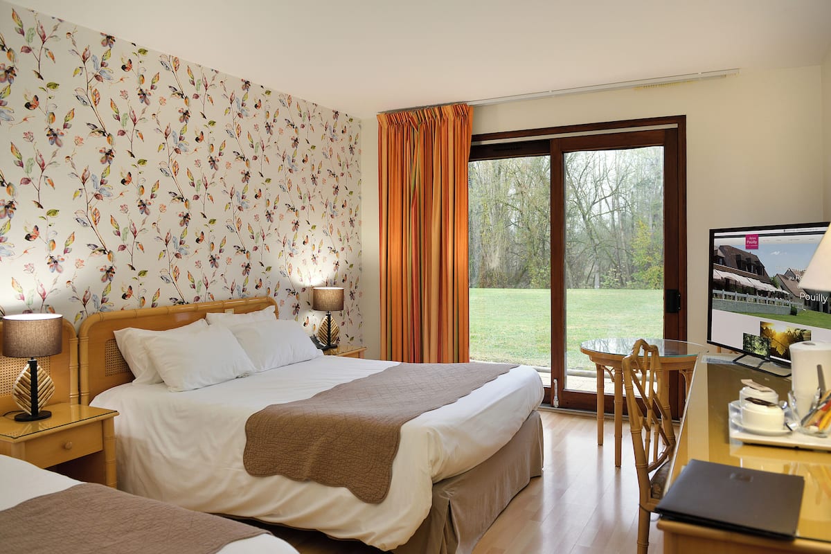 A hotel room with a large bed, a desk with a laptop, a TV, and a view of a grassy area through a sliding glass door.