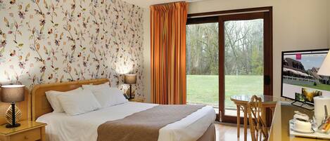 A hotel room with a large bed, a desk with a laptop, a TV, and a view of a grassy area through a sliding glass door.