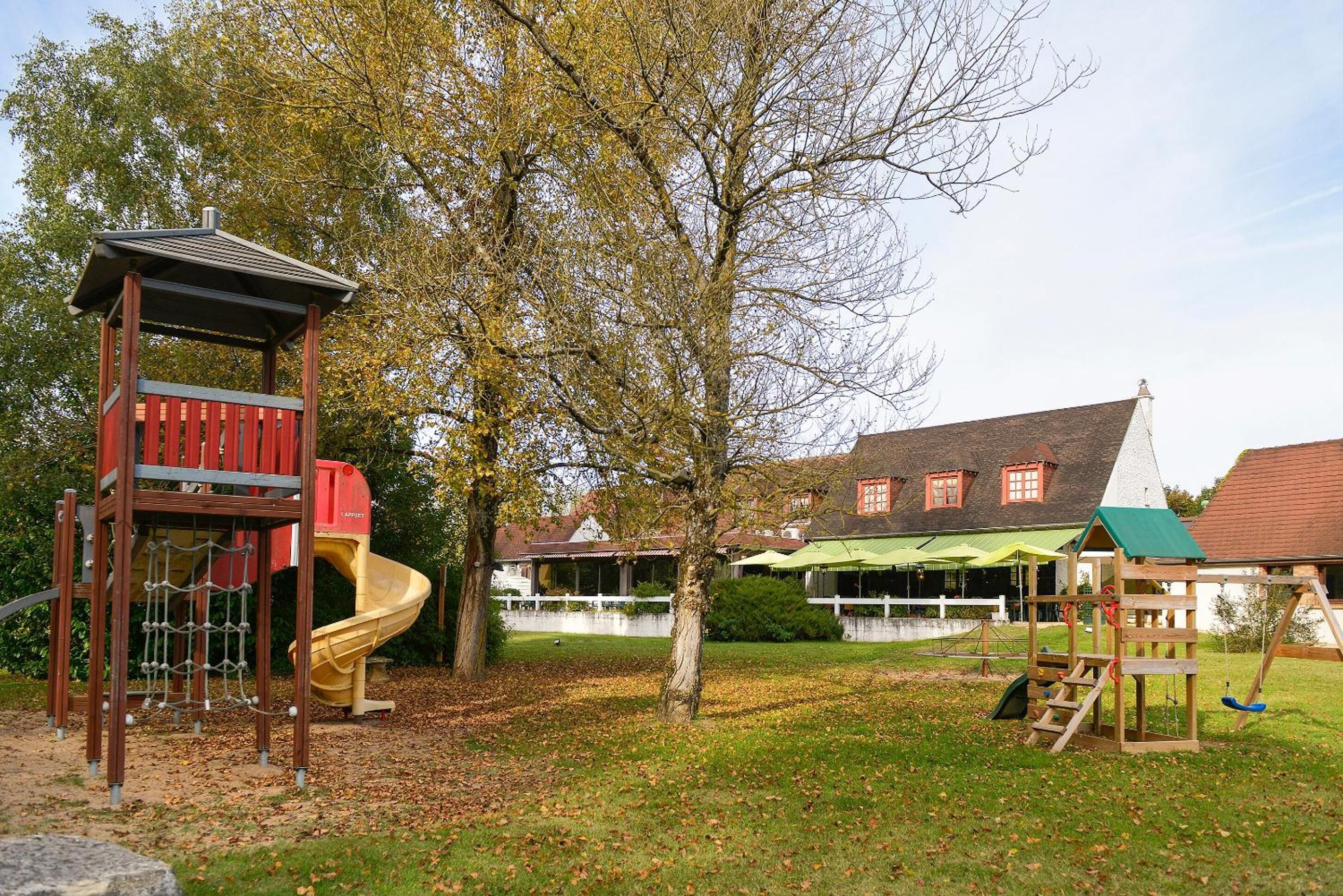 A playground with a slide and climbing structures, surrounded by trees and a building with a green roof in the background.
