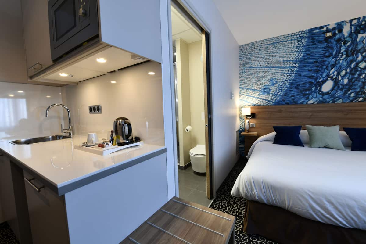 A modern hotel room with a kitchenette, a large bed with blue and green pillows, and a blue abstract wall art.