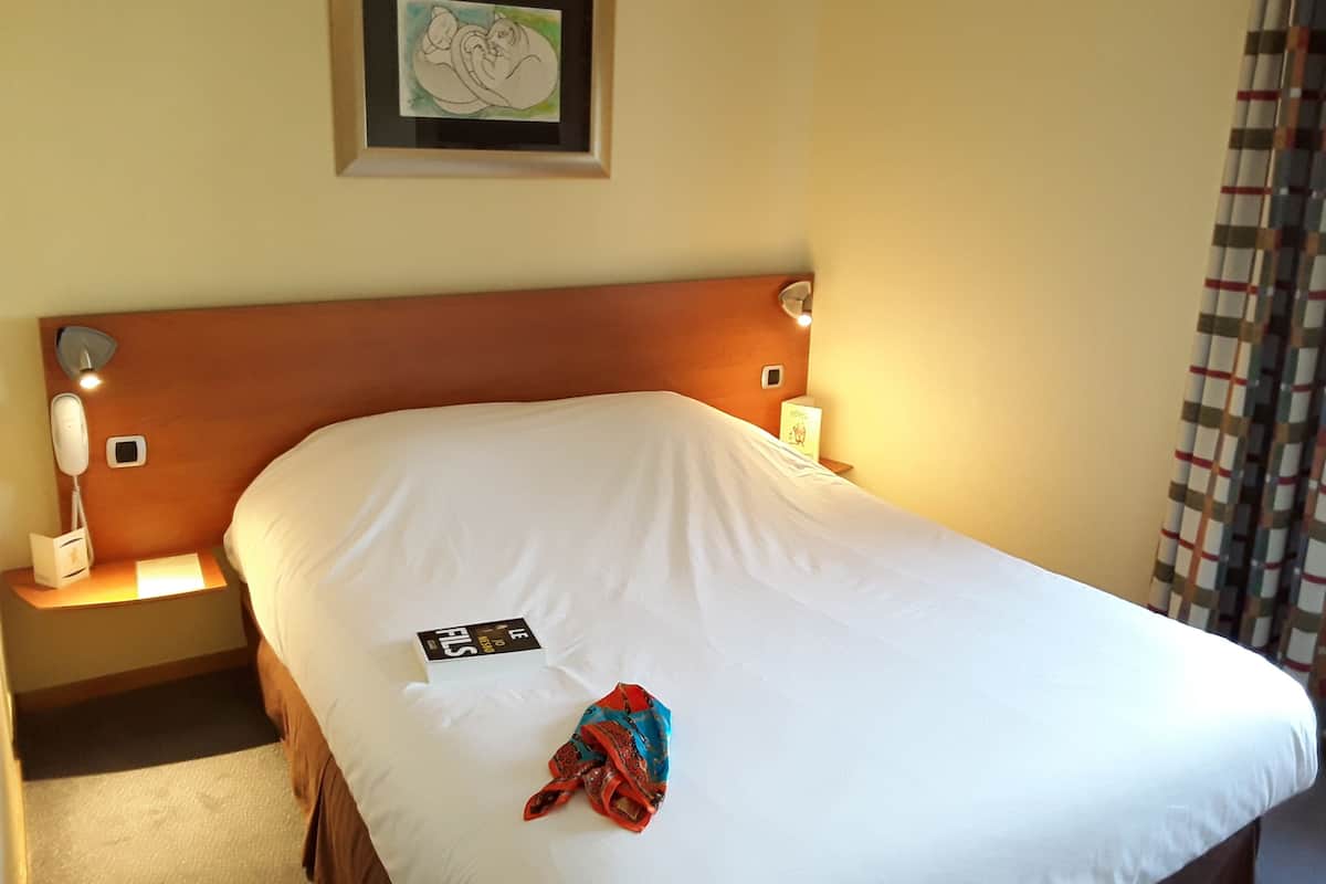 A hotel room with a wooden headboard, a white bedspread, a framed artwork on the wall, a bedside table with a lamp, and a telephone.