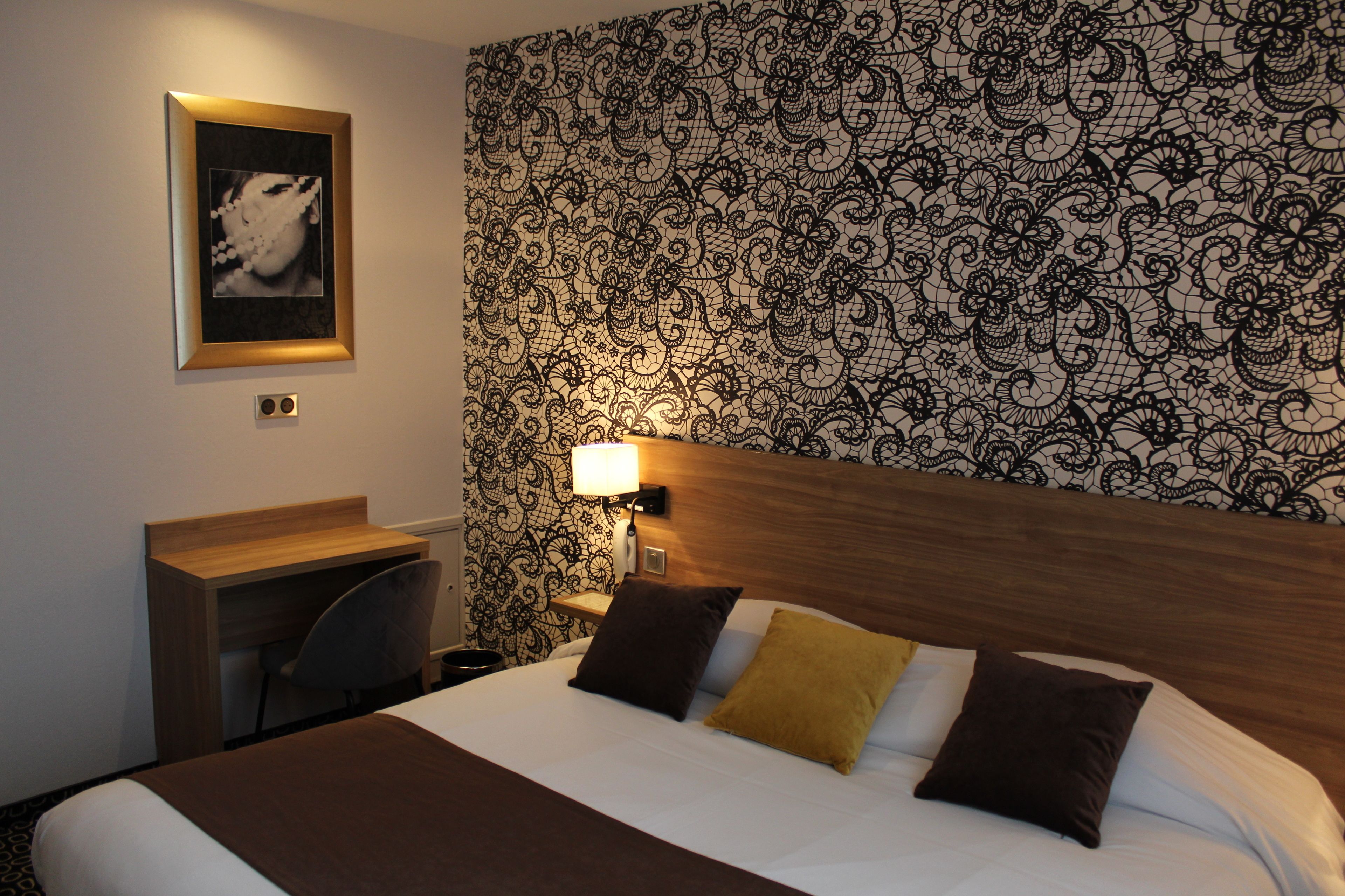 A hotel room with a bed, a desk, a chair, and a wall with a patterned wallpaper.