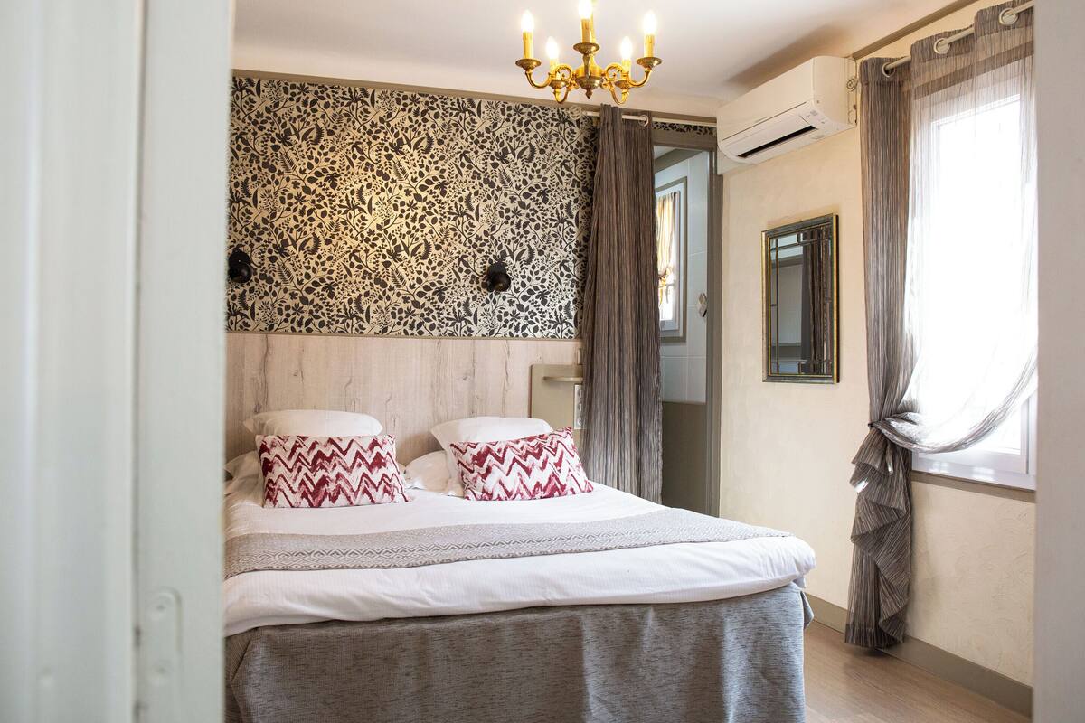 Une chambre à coucher avec un lit, un papier peint à motifs, un lustre, un appareil de climatisation, un miroir et des rideaux.