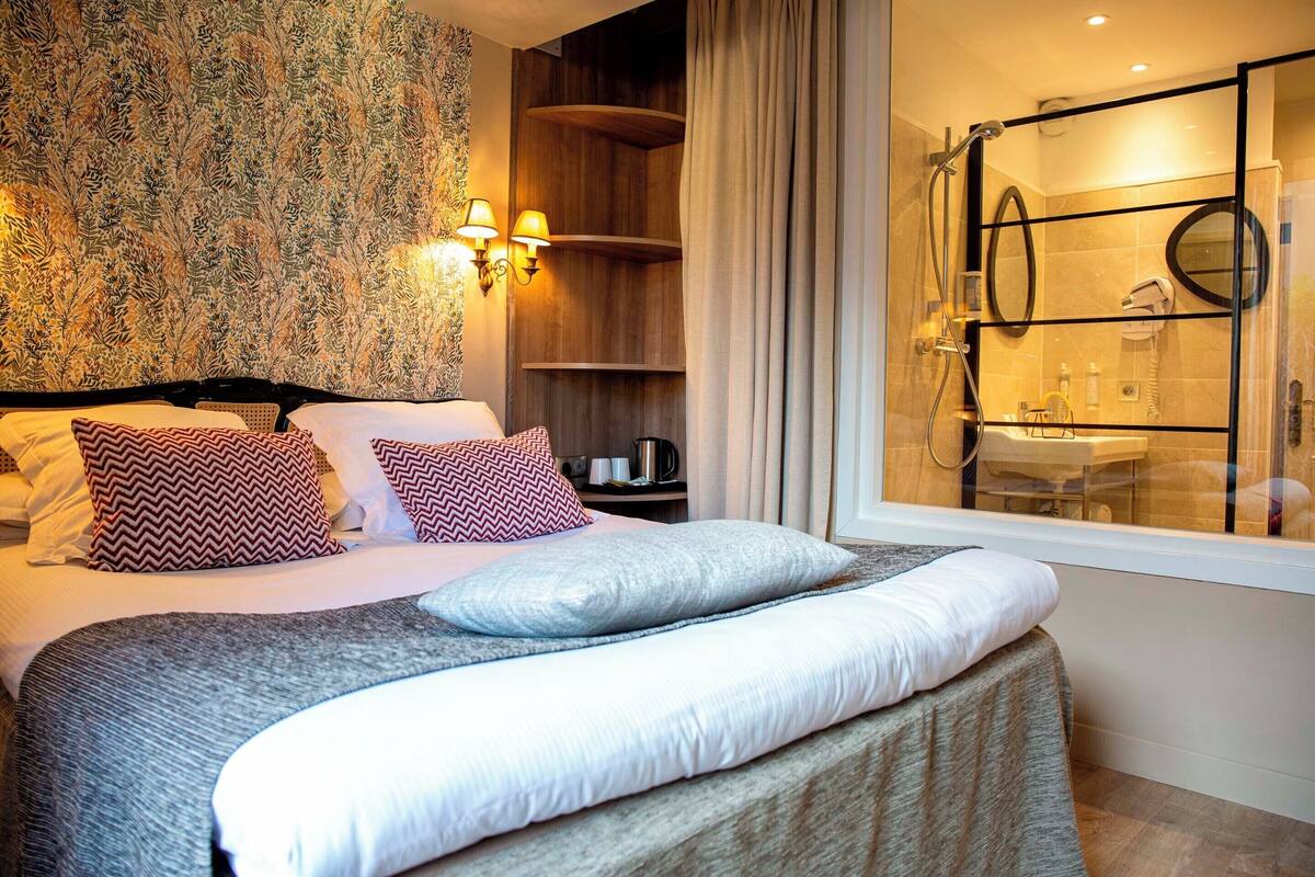 Une chambre d’hôtel avec un grand lit, un espace douche doté d’une porte en verre et un mur recouvert d’un papier peint à motifs floraux.