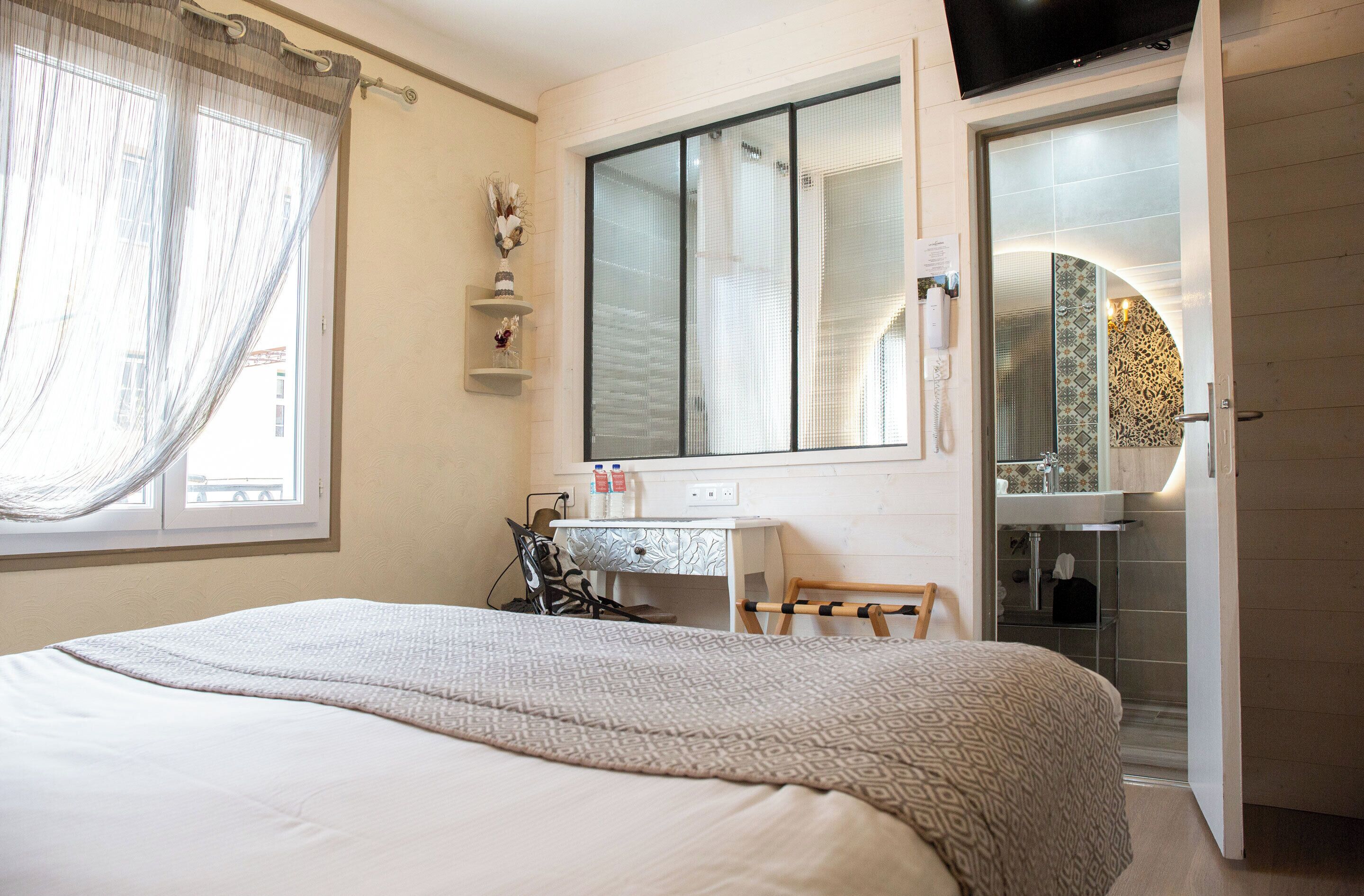 A bedroom with a bed, a desk, a chair, a mirror, and a bathroom visible through an open door.