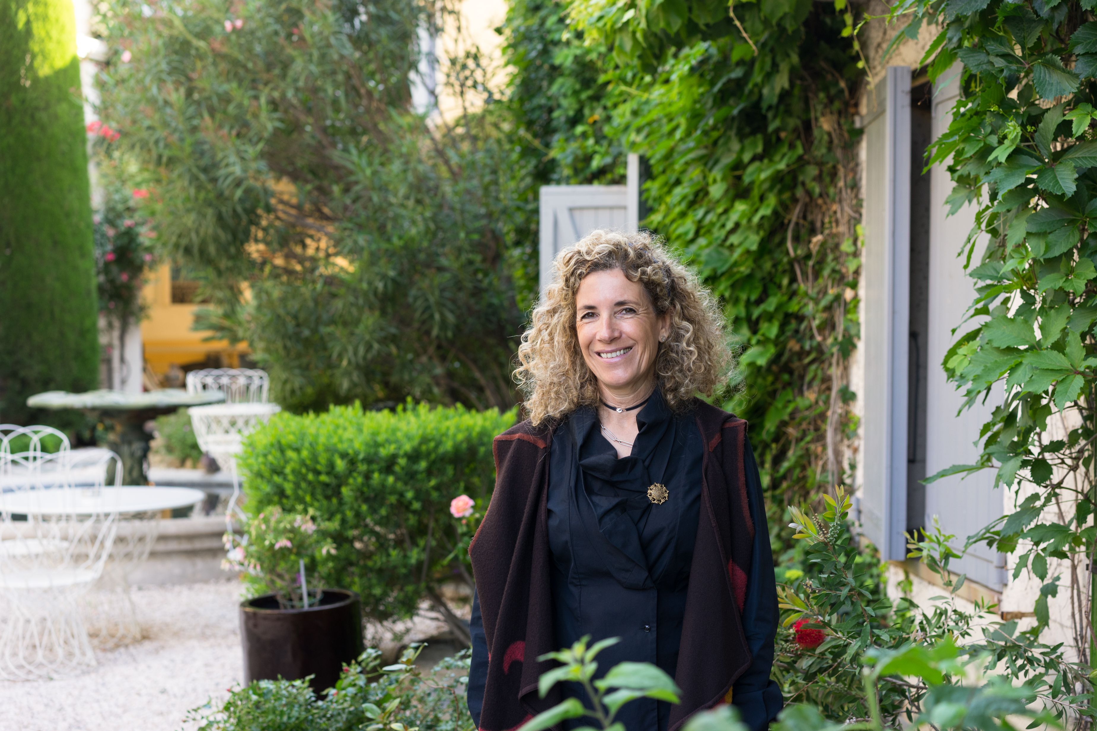 Una persona de pie en un jardín con una mesa y sillas blancas, rodeada de follaje y un edificio.