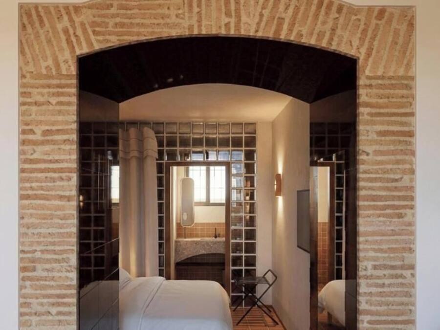 A hotel room with a brick archway, a bed, a desk, and a bookshelf.
