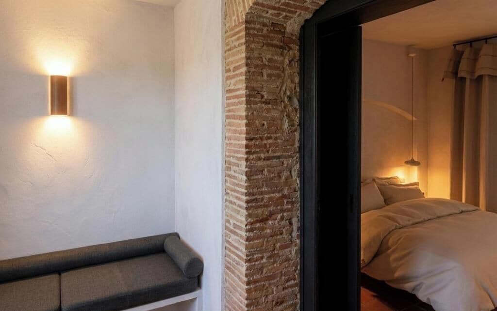 A hotel room with a bed, a bench, a brick wall, and a wall-mounted light fixture.