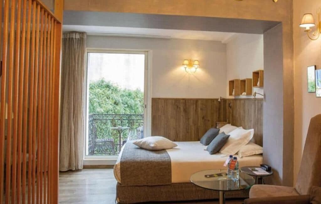 Habitación de hotel con cama, una mesita, una silla, balcón con vistas a la vegetación y cabecero de madera.