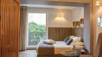 Habitación de hotel con cama, una mesita, una silla, balcón con vistas a la vegetación y cabecero de madera.