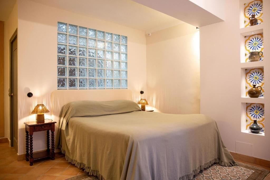 Una camera da letto con un'ampia finestra, due lampade ai comodini, un letto con copriletto color beige e mensole decorative a parete.