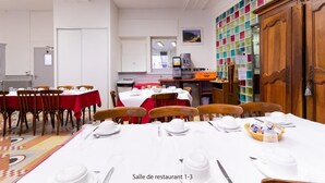 A dining area with tables set for a meal, a kitchen area with appliances, and a wall of colorful storage bins. - Croix de Malte (Lourdes)
