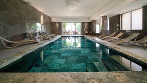 A modern indoor swimming pool with a clear water surface, surrounded by lounge chairs and a stone-tiled floor.