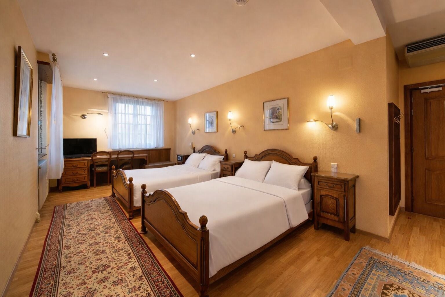 A hotel room with two wooden beds, a desk with a chair, a television, and a large window with curtains.