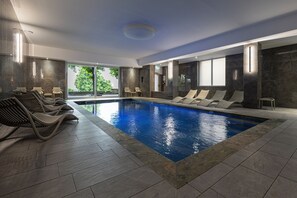 A modern indoor swimming pool with lounge chairs and a view of greenery outside.