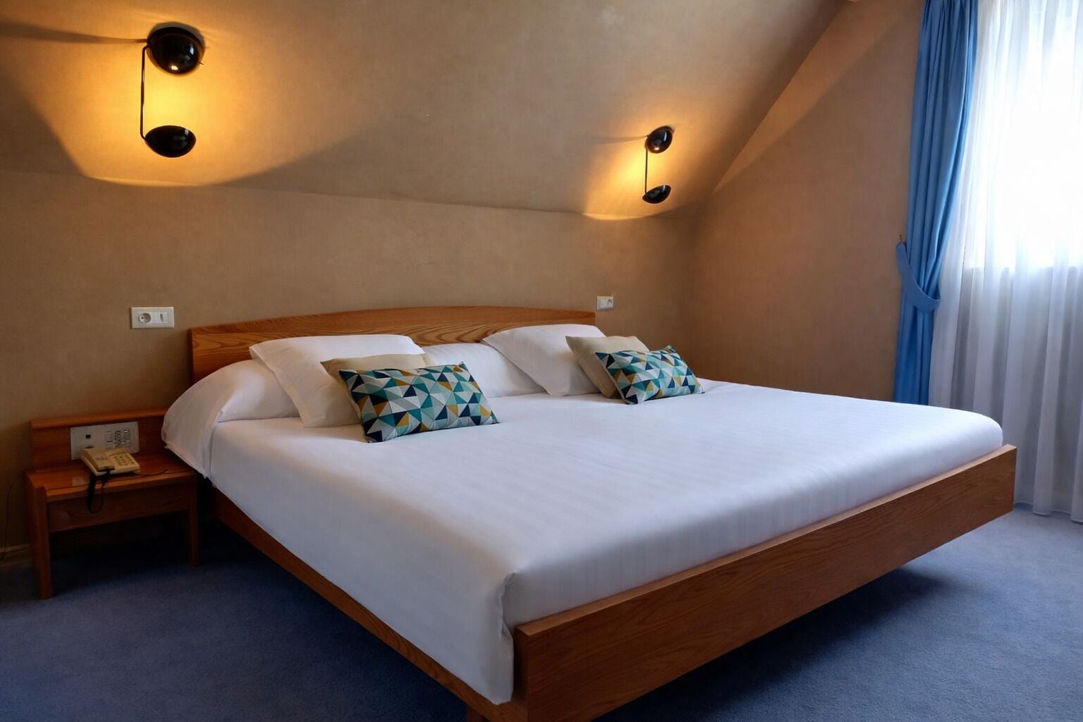 Una cama grande con estructura de madera, dos mesitas de noche y un teléfono sobre la de la izquierda.