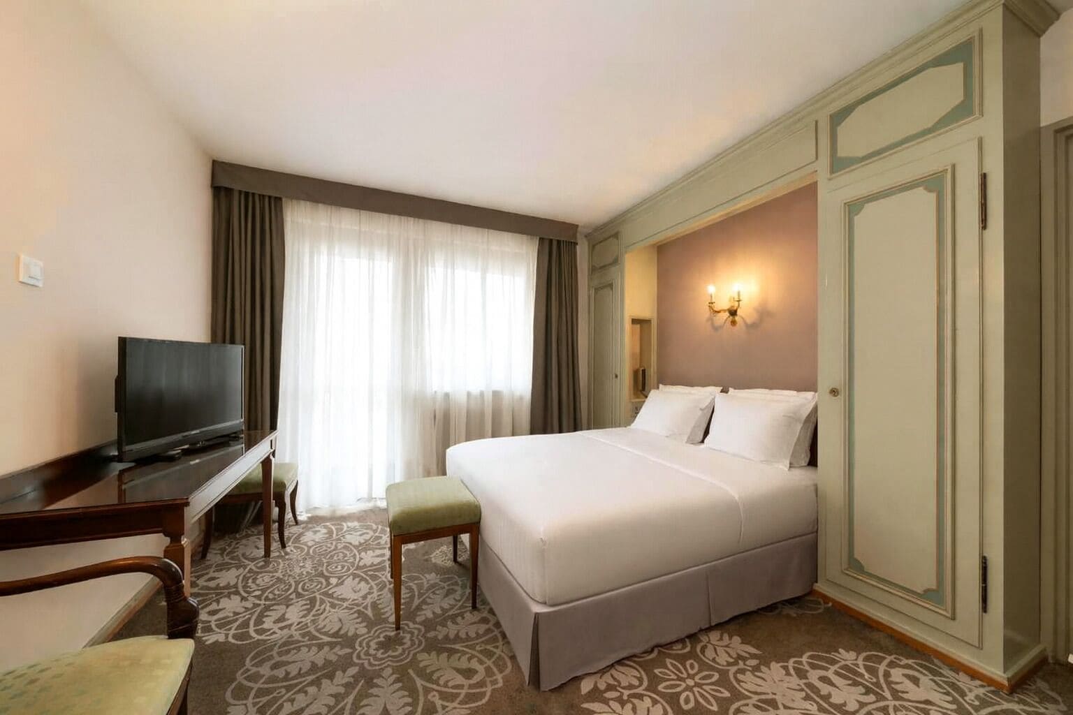 A hotel room with a bed, a desk with a TV, a chair, and a wardrobe.