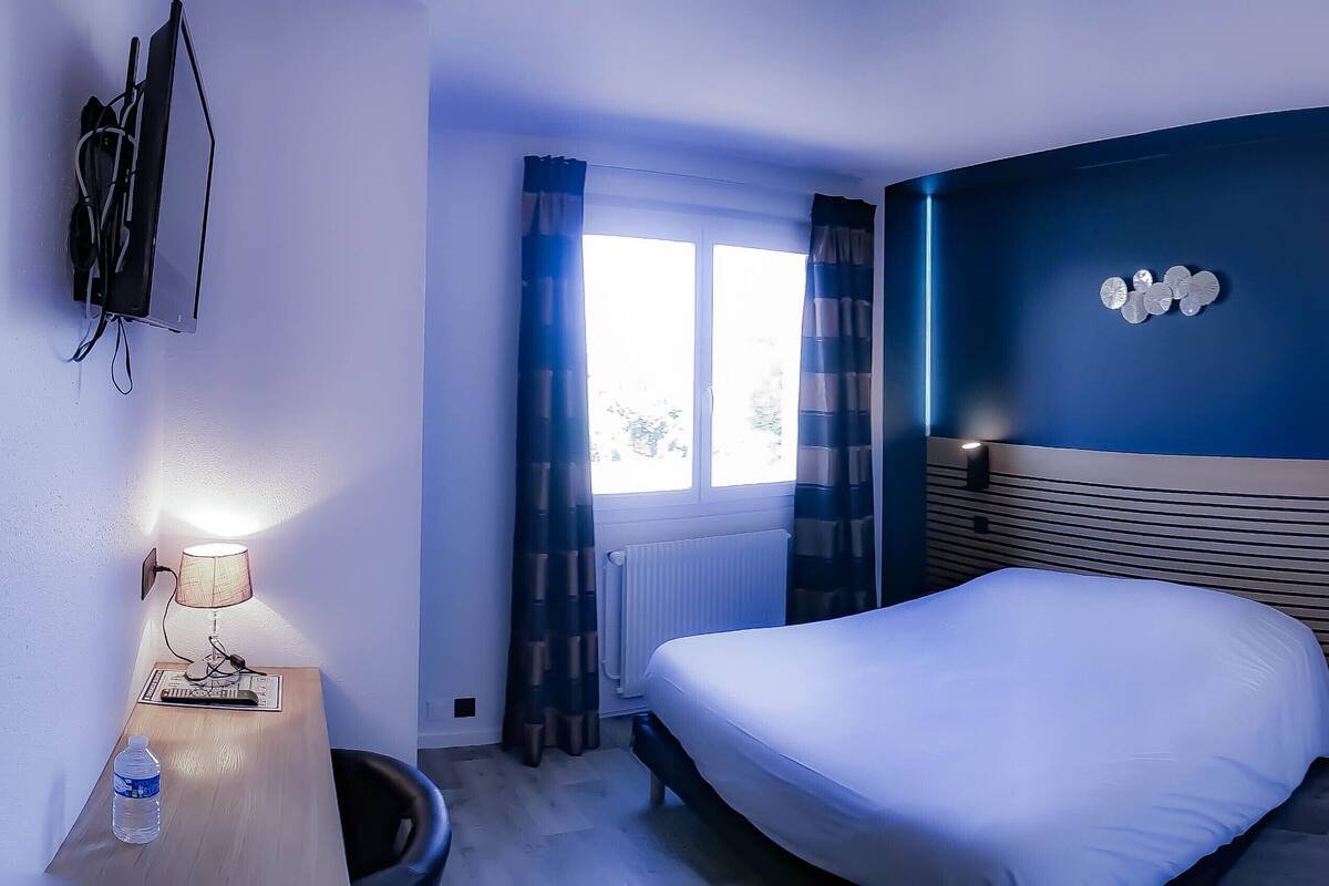 Une chambre d’hôtel avec un lit, un bureau doté d’une lampe, une bouteille d’eau et une fenêtre avec des rideaux.