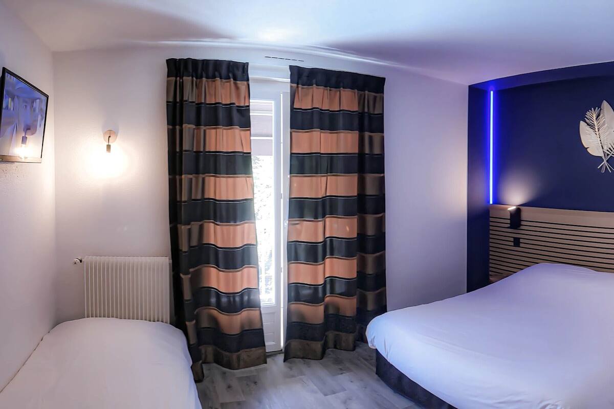 Une chambre d’hôtel avec deux lits, une grande fenêtre avec des rideaux, un luminaire fixé au mur et un élément décoratif représentant des feuilles.
