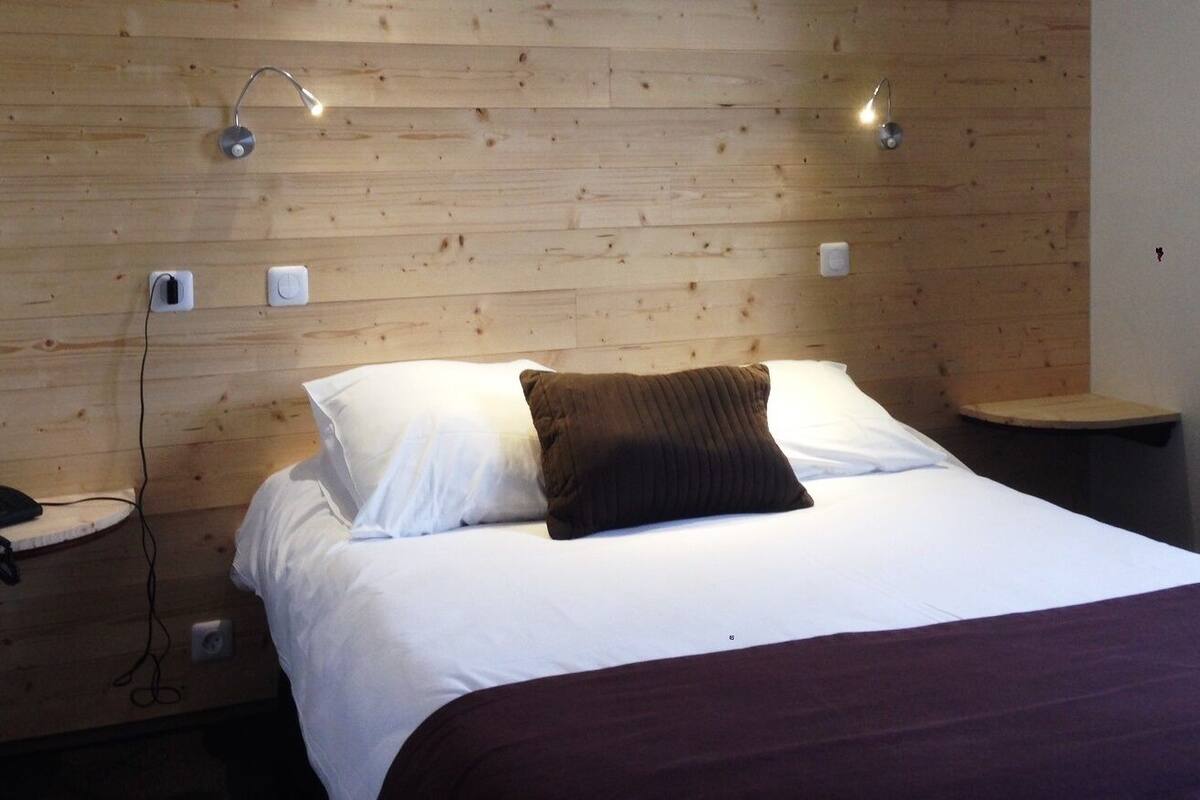 Une chambre d’hôtel avec une tête de lit en bois, un lit recouvert de draps blancs, un coussin décoratif marron, des tables de chevet avec des lampes et un téléphone sur l’une des tables.