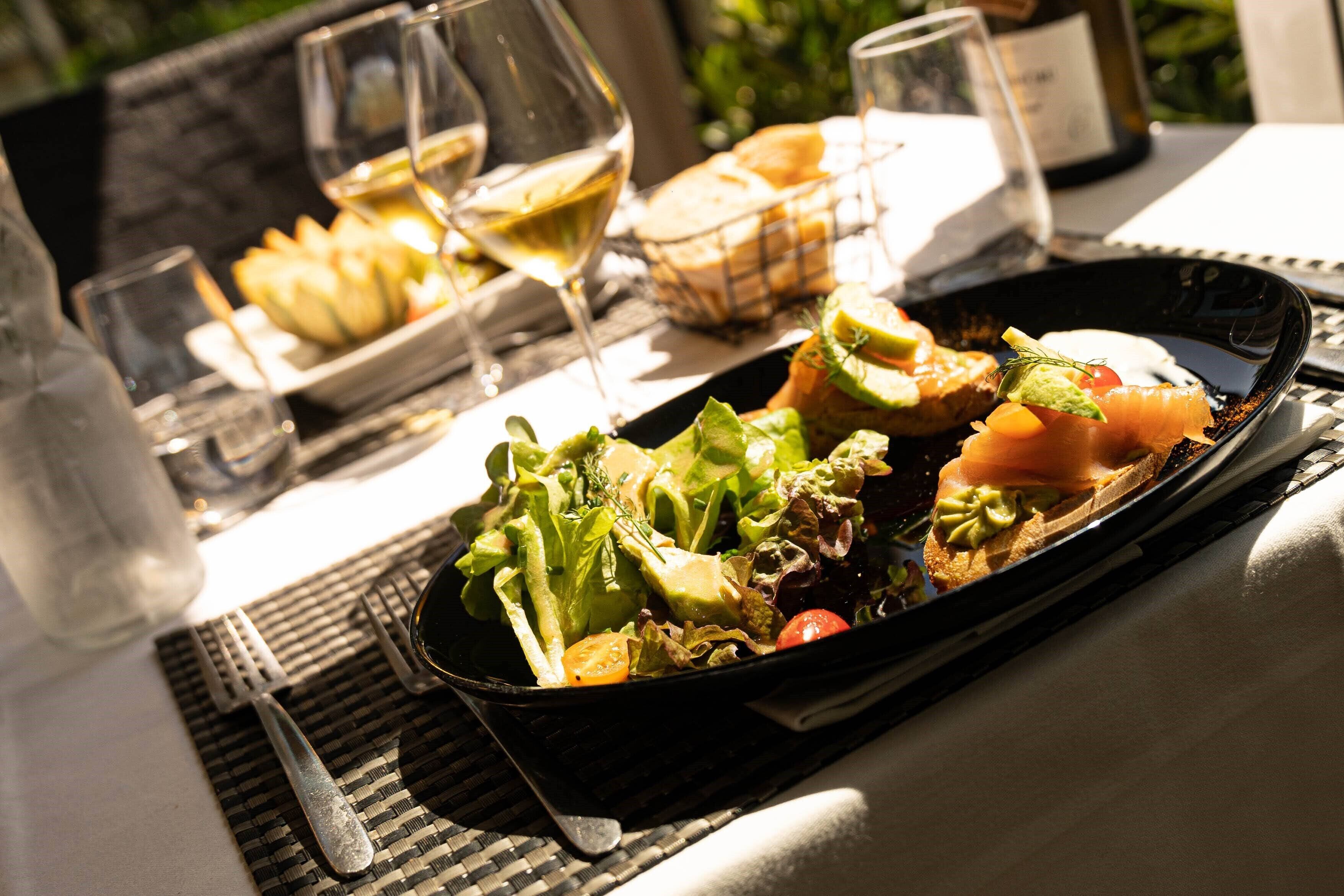 Une table à manger avec une assiette noire contenant une salade, une tranche de saumon et un morceau de pain. On y trouve également deux verres de vin blanc et une bouteille de vin.