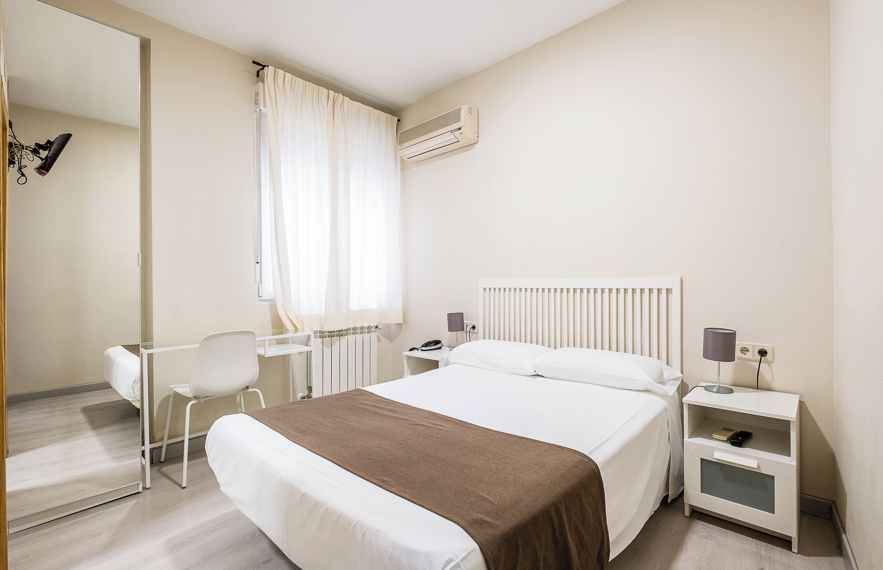 A hotel room with a bed, bedside tables, a desk, a chair, a mirror, and a radiator.
