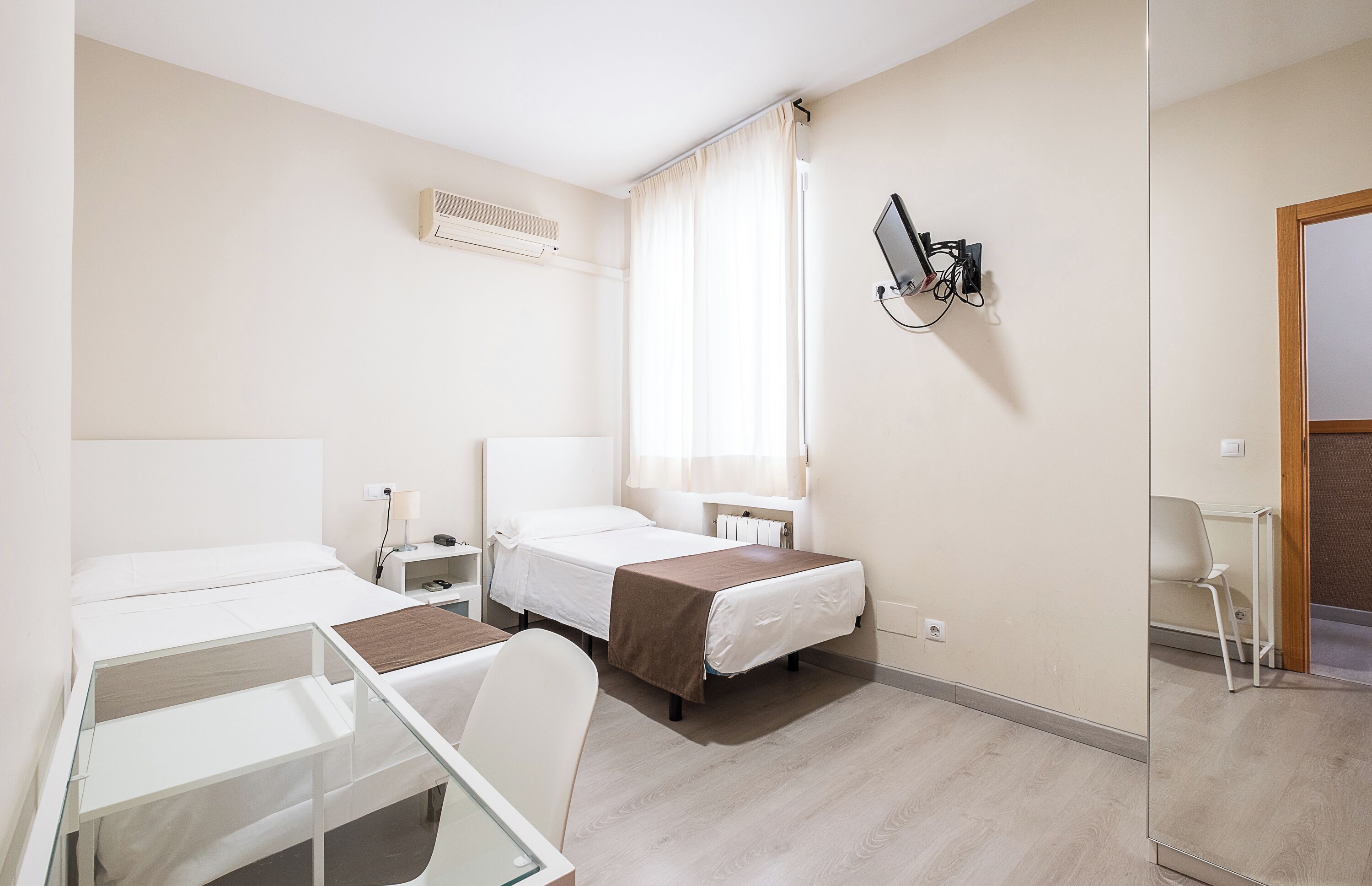 A hotel room with two single beds, a bedside table with a lamp, a small desk, and a chair.