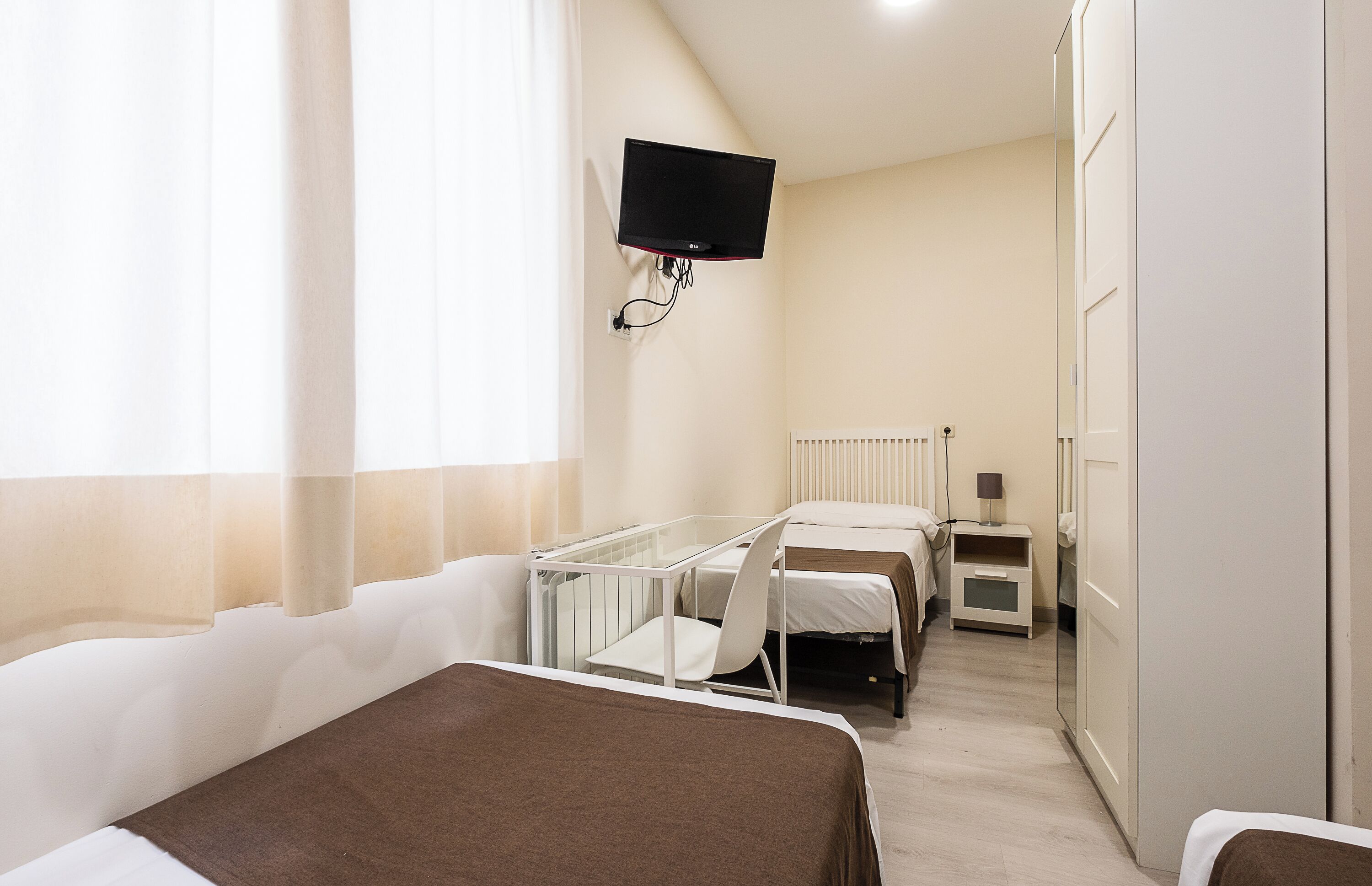 A hotel room with a bed, a bedside table, a lamp, a TV mounted on the wall, and a window with curtains.