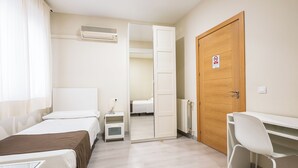 A hotel room with two beds, a desk, a chair, a radiator, and a door with a no-smoking sign. - Hostal El Pilar (Madrid)