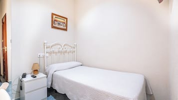 Een kleine eenpersoonskamer met een witte metalen hoofdeinde, een nachtkastje met een lamp, en een aan de muur bevestigd schilderij.