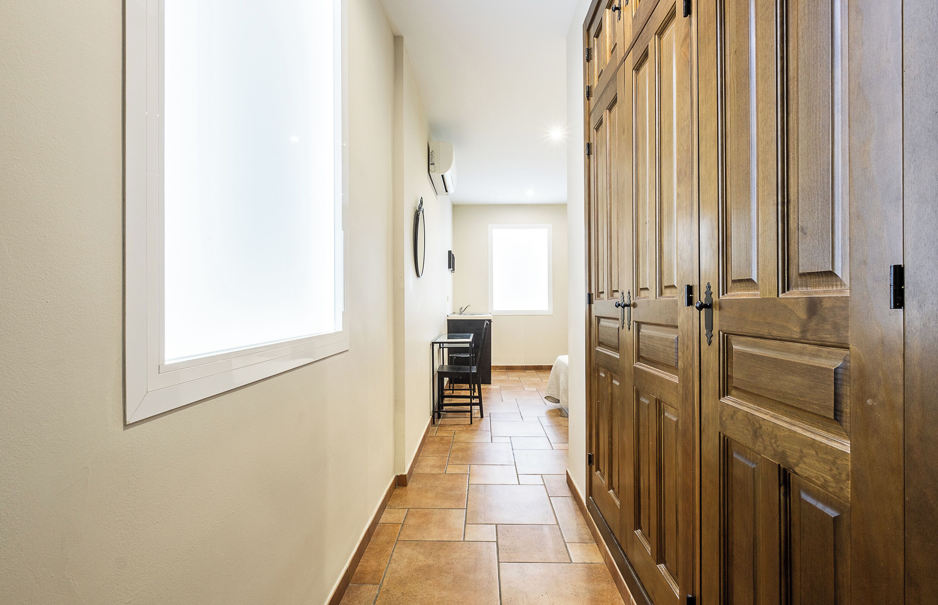 A hallway with a large window, a wooden wardrobe, and a desk with chairs.