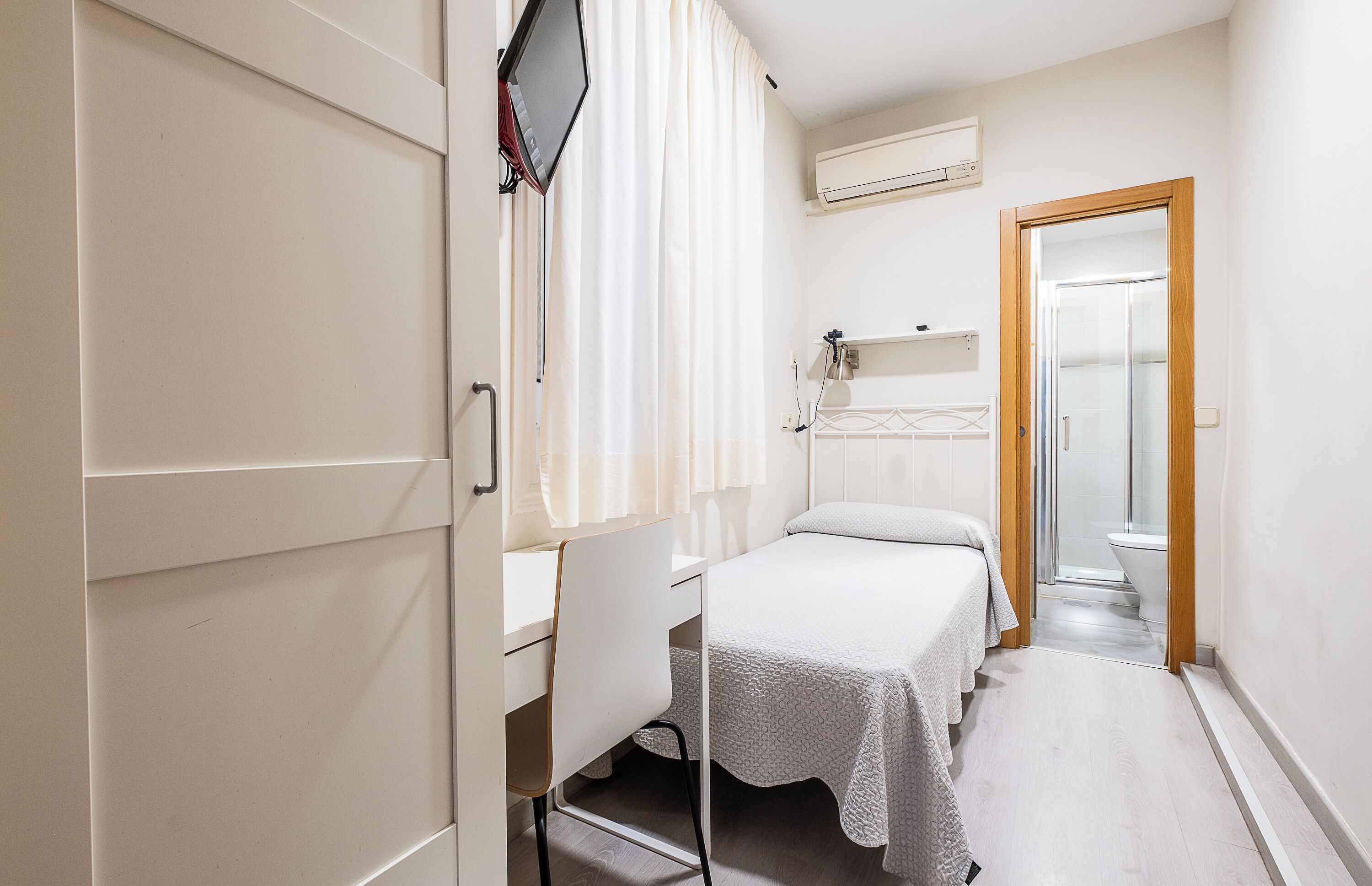 Una habitación de hotel pequeña y estrecha con una cama individual, una mesita de noche, un espejo y un rincón de ducha.
