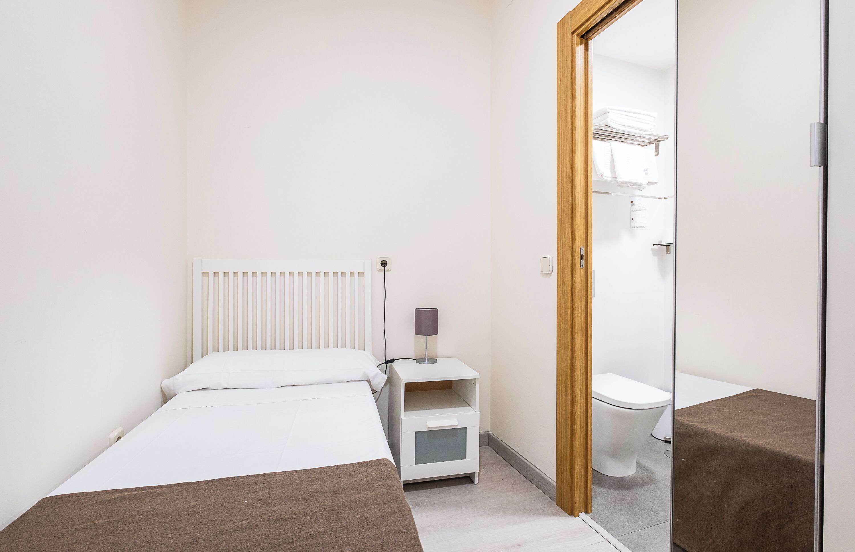 A small, single-bed room with a radiator, a bedside table with a lamp, and a toilet visible through an open door.