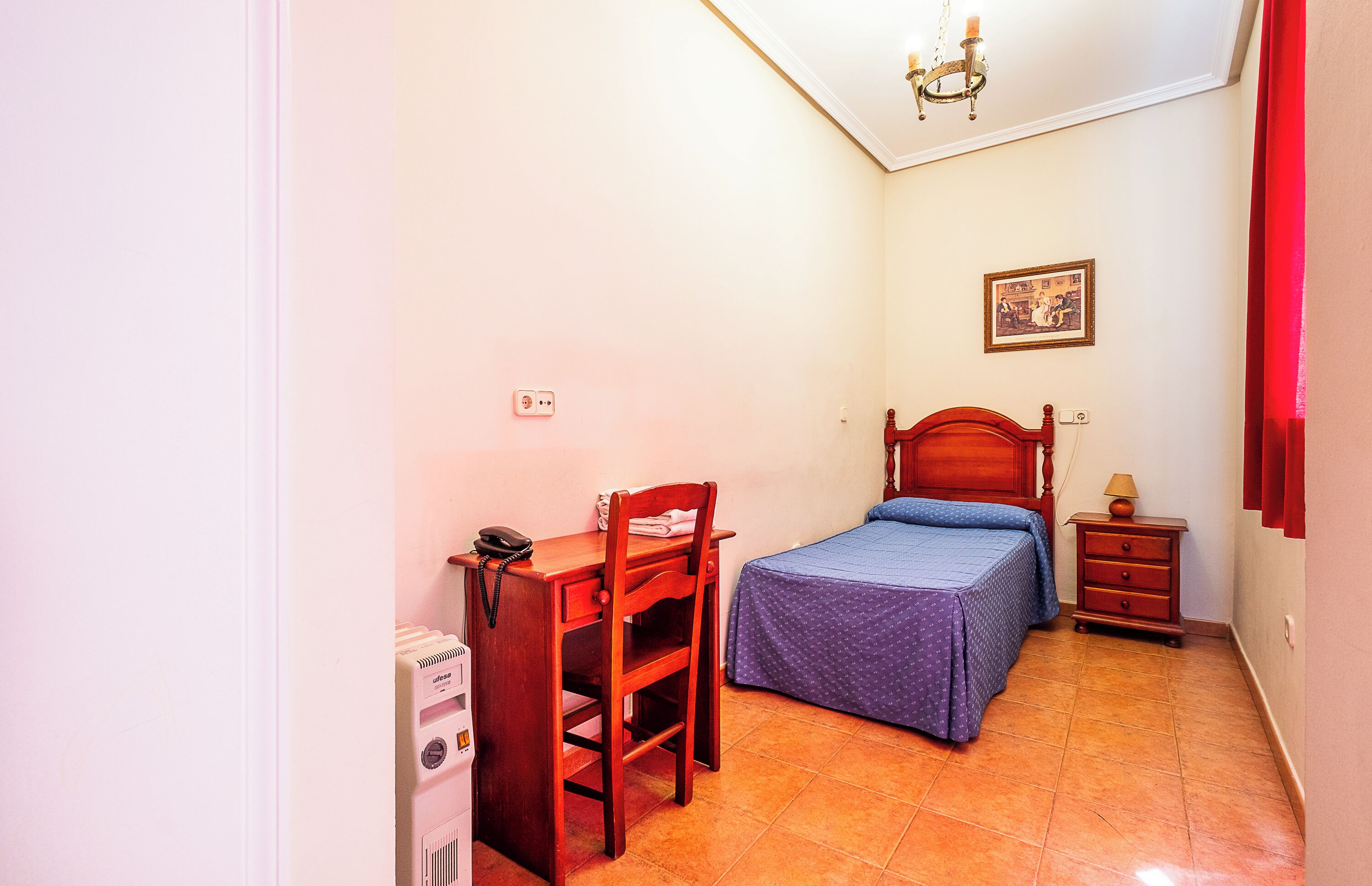Een kleine slaapkamer met een enkel bed, een nachtkastje, een bureau met stoel, een telefoon aan de muur en een plafondlamp.
