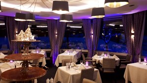 Un restaurante con mesas redondas, lámparas colgantes negras y vista a un paisaje urbano nocturno.