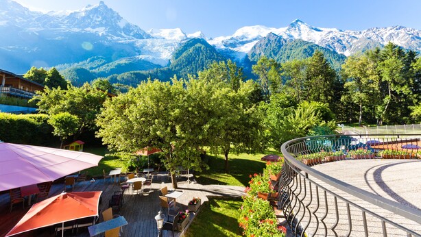 Una terraza con mobiliario exterior, rodeada de abundante vegetación y vistas a montañas nevadas.
