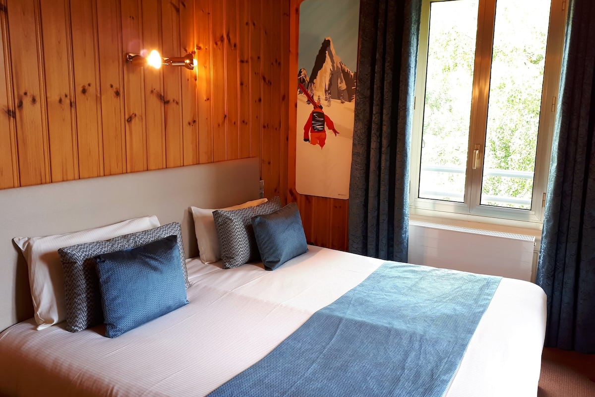 Ein Hotelzimmer mit einer holzgetäfelten Wand, einem Bett mit weißblauer Bettdecke und einem Gemälde von einer Berglandschaft.