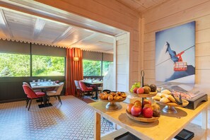 A dining area with a table set for two, a fruit bowl, and a buffet with pastries and meats.