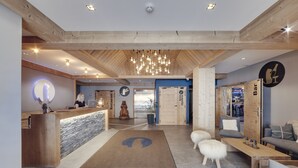 Un moderno vestíbulo de hotel con un mostrador de recepción de madera, una alfombra grande con un logotipo, una zona de estar con sofá y reposapiés, y un letrero de bar.