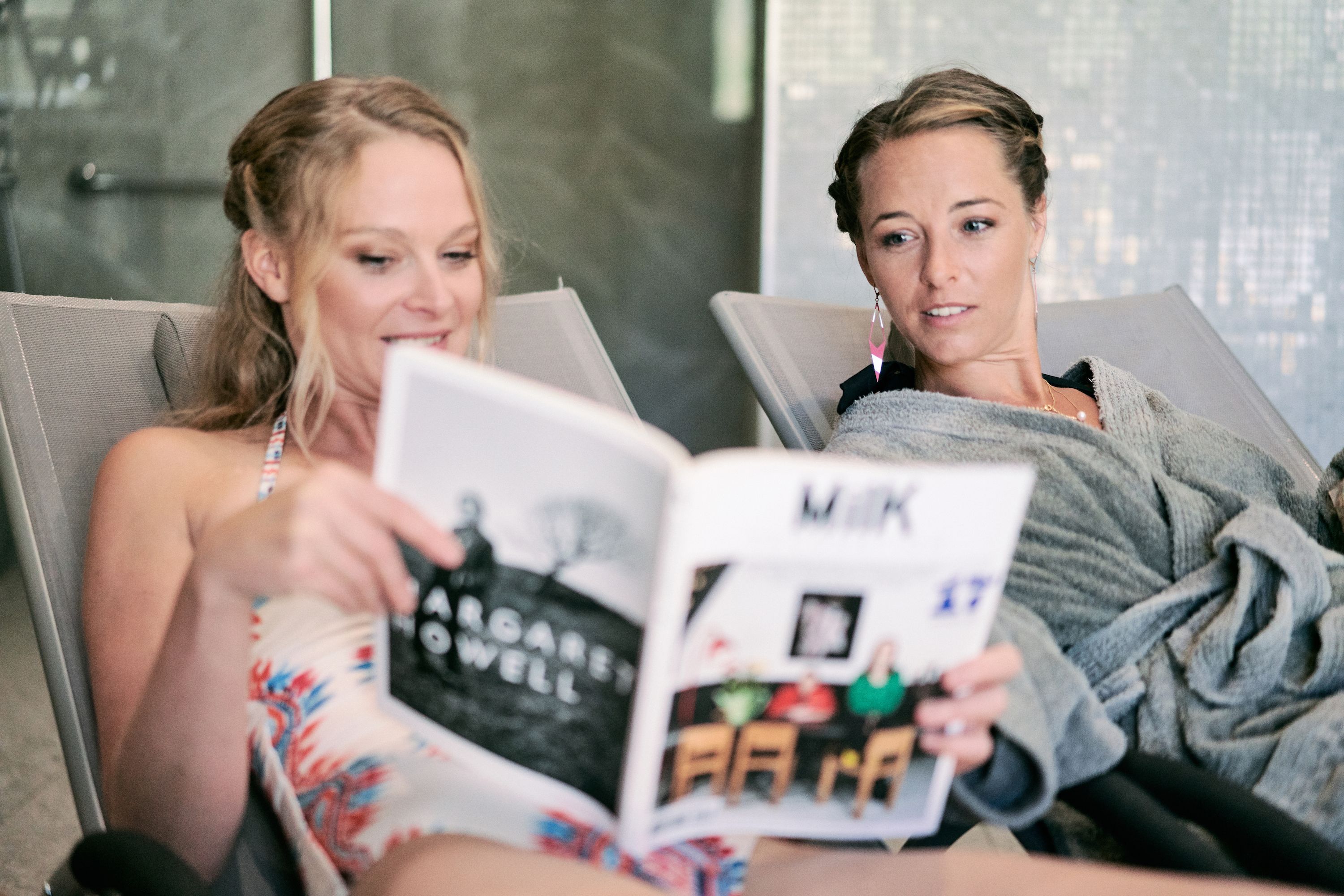 Two individuals seated in a modern lounge area with a cityscape view, reading a magazine.