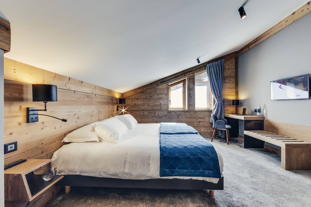 A modern bedroom with a wooden sloped ceiling, a bed with a blue and white duvet, a desk with a chair, and a TV on the wall.