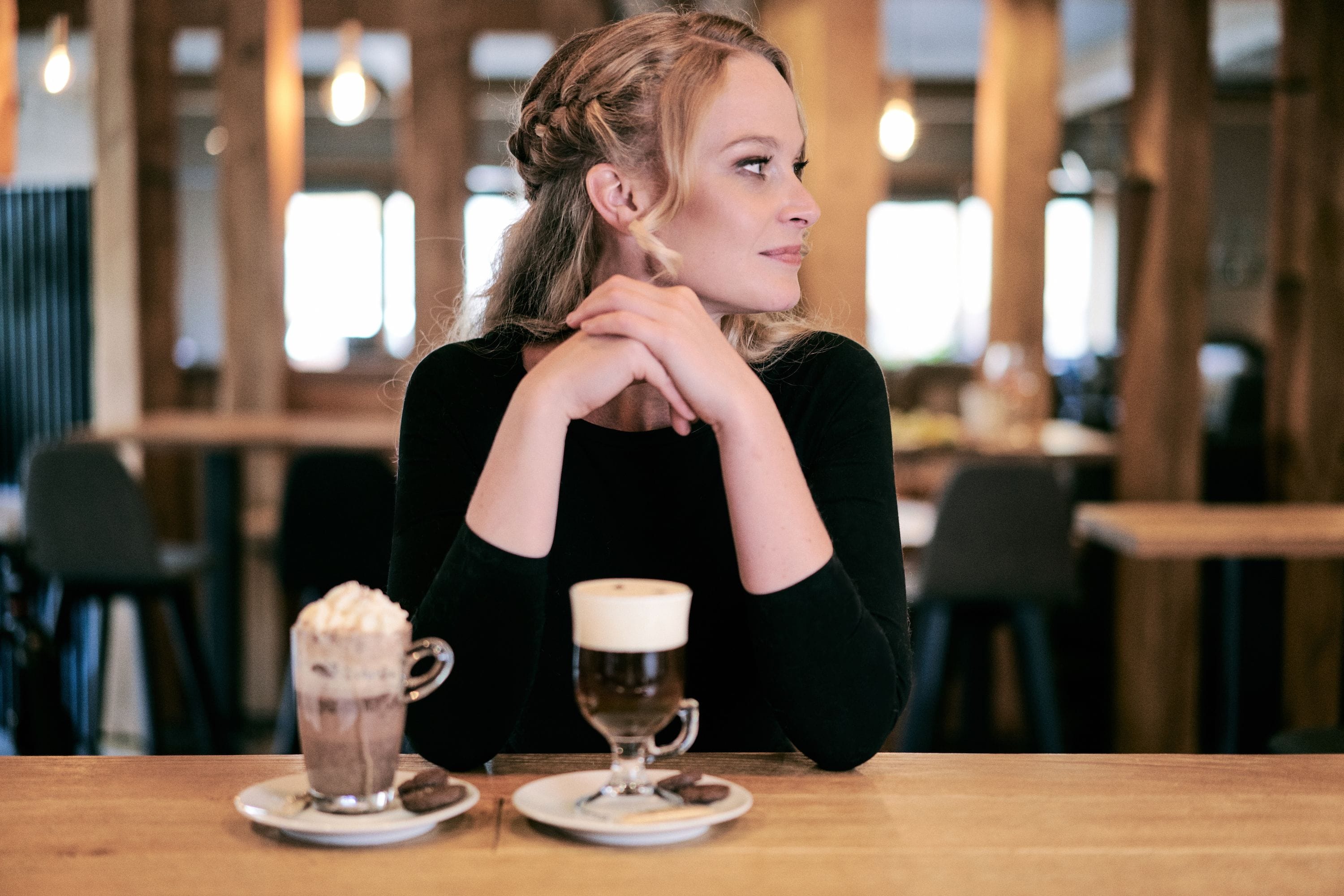 Een persoon zit aan een tafel met twee drankjes in een café met houten meubels en een modern interieur.