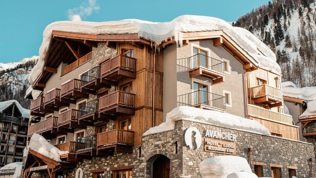 A multi-story hotel with wooden balconies and a stone facade, surrounded by snow-covered mountains.