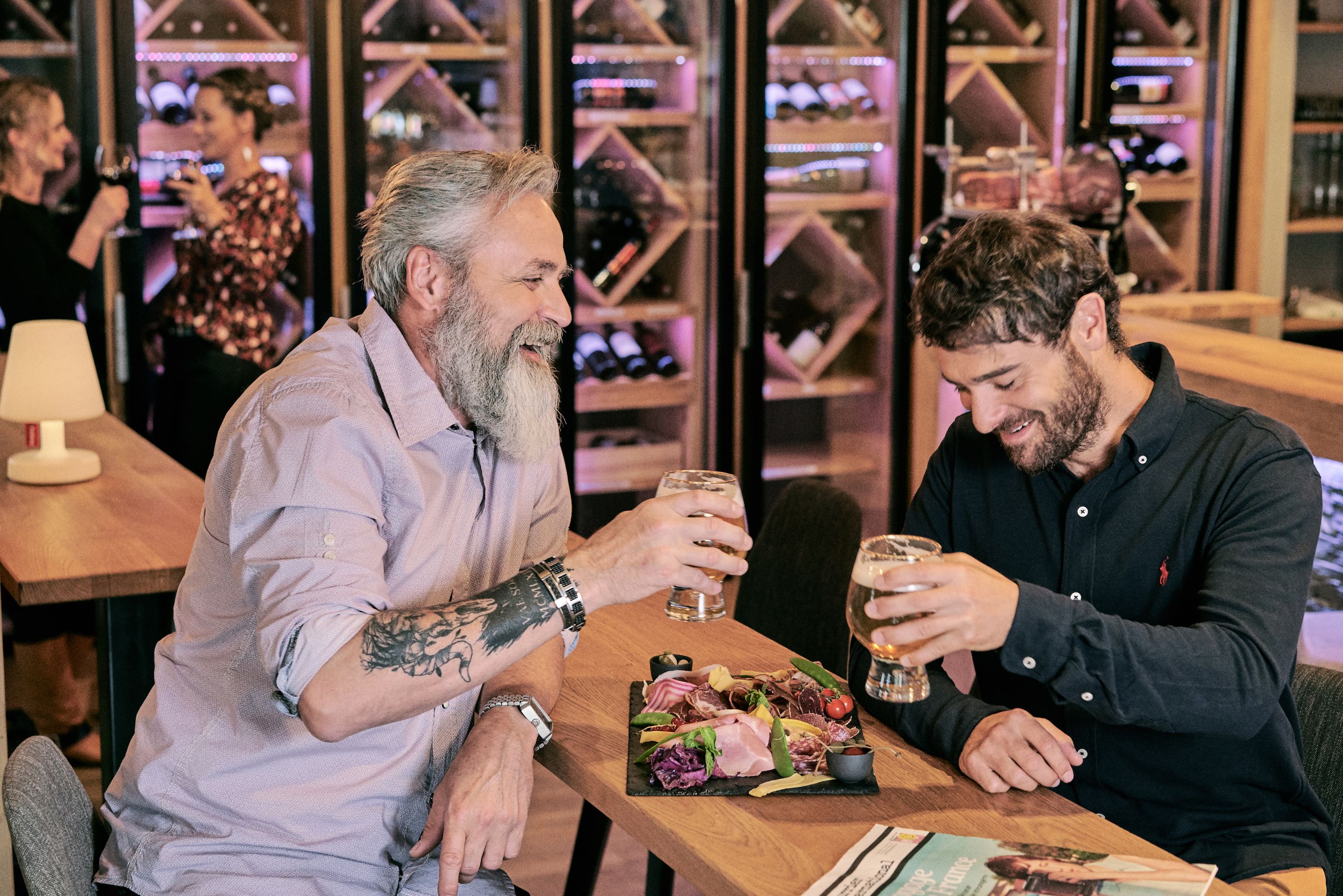 Two men seated at a table with a variety of cheeses and meats, glasses of beer, and a magazine.