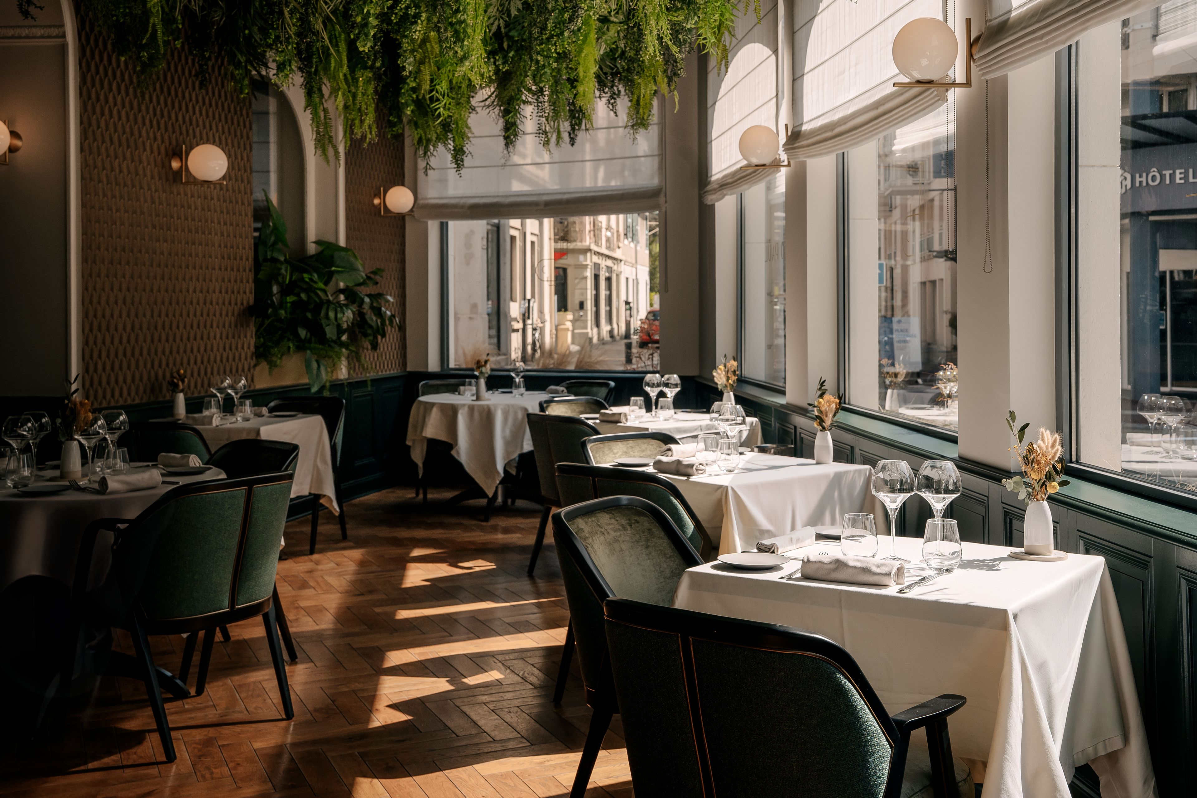 A restaurant interior with tables set for dining, hanging plants, and a view of the street through large windows.