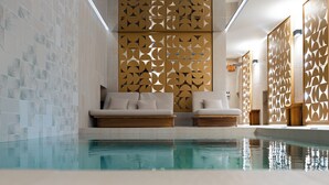 Un espace piscine intérieur moderne, doté d’une paroi en verre, de deux chaises longues et d’un mur décoratif orné de motifs géométriques.