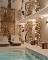 Un espace piscine intérieur moderne, avec un mur aux motifs géométriques, un fauteuil de relaxation et une personne portant un peignoir.
