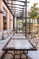 Un área de estar al aire libre techada, con una mesa larga y sillas, un sofá con cojines y una estructura de pérgola.
